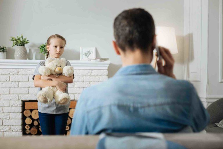 Vater telefoniert am Sofa und Tochter steht mit Kiuscheltier davor